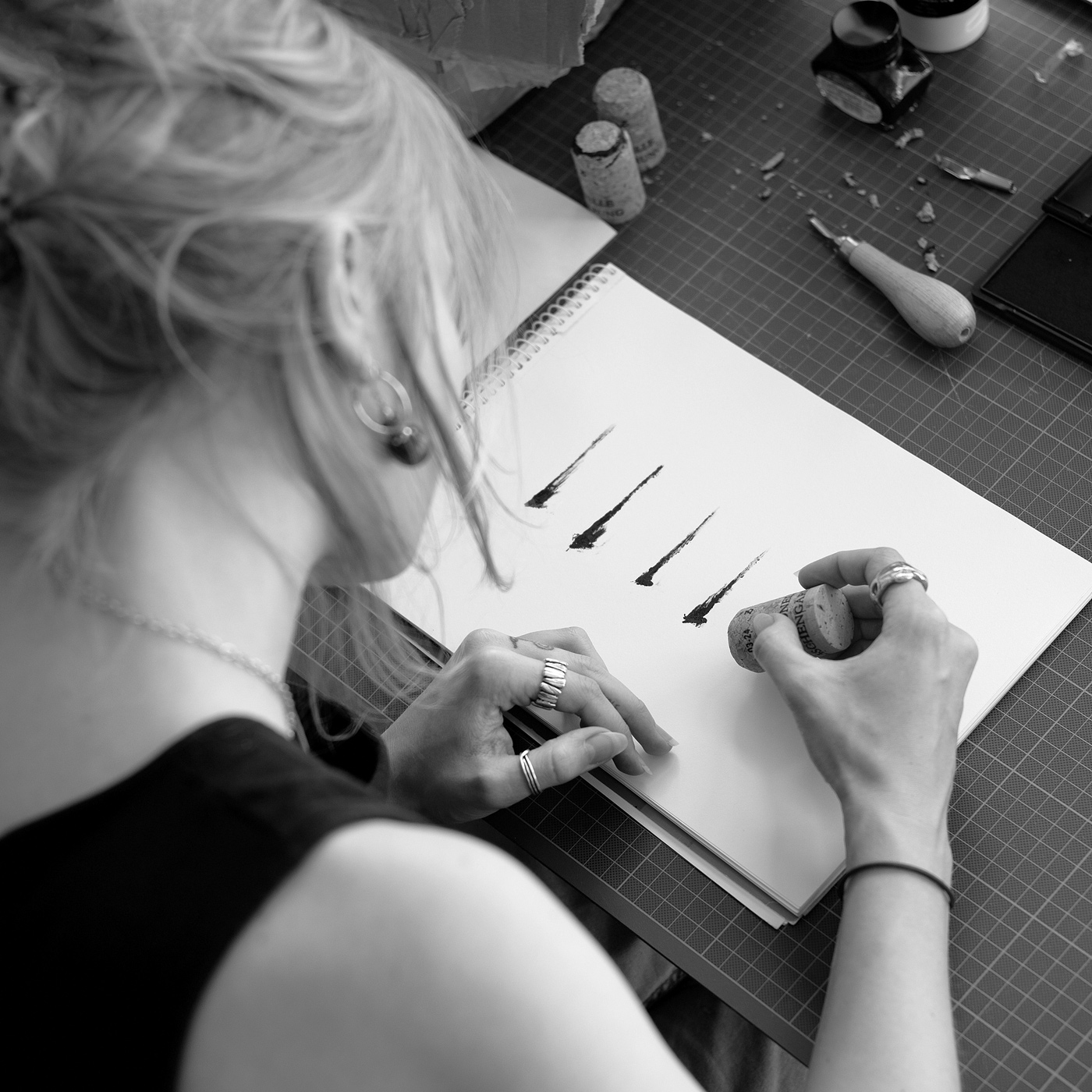 Person druckt mit einem Korken-Stempel mehrere schwarze Striche in ein Skizzenbuch; Werkzeuge liegen am Tisch.